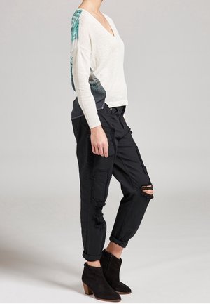 Maglione bianco a V con foglie di palma verdi sui lati, abbinato a pantaloni cargo neri con strappi e tasche, e ankle boots neri.