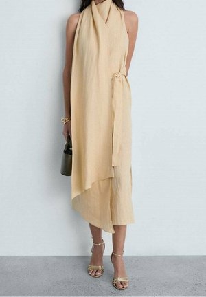 Kobieta ubrana w beżową sukienkę na ramiączkach z wiązaniem z boku, trzymająca małą ciemną torebkę, ubrana w złote sandały na obcasie i bransoletkę.