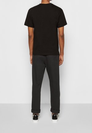 Osoba stojąca tyłem, ubrana w jednolitą czarną koszulkę, ciemne podwinięte spodnie oraz czarno-białe trampki na jasnoszarej podłodze.