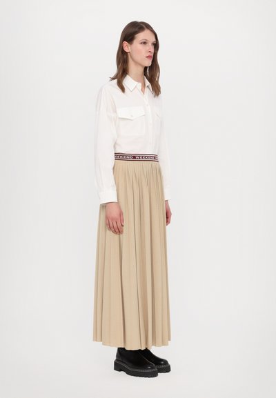 Chemise blanche à boutons avec deux poches poitrine, associée à une jupe maxi plissée beige avec une taille élastique bordeaux. Bottes noires à semelle épaisse.