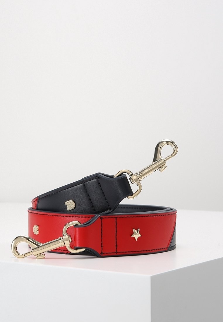 Bracelet en cuir rouge et noir avec fermoirs dorés et petits clous en métal en forme d’étoile et de visage, enroulé sur une surface blanche.