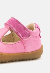 Vaaleanpunaiset nahkasaappaat, joissa on pyöristetty kärki, tarranauhakiinnitys ja vaaleanruskea kumipohja. Korostaa teksturoitua pintaa ja hienovaraisia ompeluyksityiskohtia.