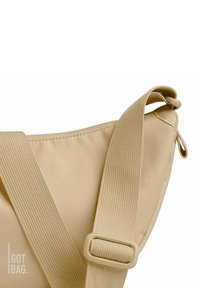 Bolso de hombro beige fabricado con un material duradero, con una correa ancha y una superficie texturizada. Presenta un broche y detalles de costura.