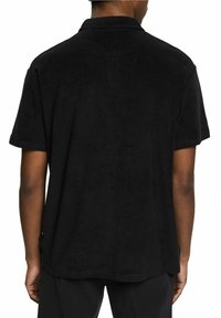 Personne portant un polo noir à manches courtes et un pantalon noir, vu de dos sur un fond blanc uni.