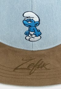 Blaue Denim-Kappe mit einem braunen Wildlederschirm, verziert mit einem gestickten blauen Charakter mit weißen Akzenten. "Zefix" ist auf dem Schirm eingeprägt.