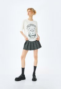 Hvid t-shirt med et bjørne grafik og teksten "CALIFORNIA", parret med en grå ternet plisseret nederdel. Modellen bærer sorte platform sko og grå sokker.