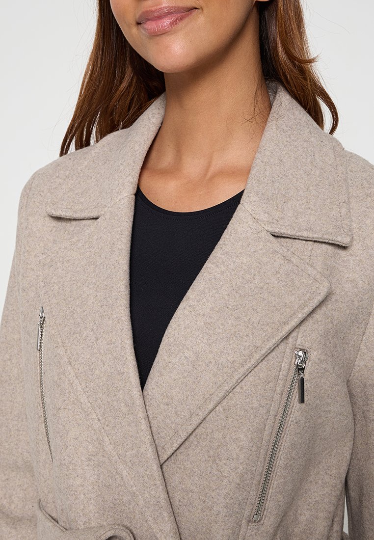 Frau, die einen beigen Wollmantel mit breiten Revers und vertikalen silbernen Reißverschlusstaschen über einem schwarzen Oberteil trägt und leicht lächelt.