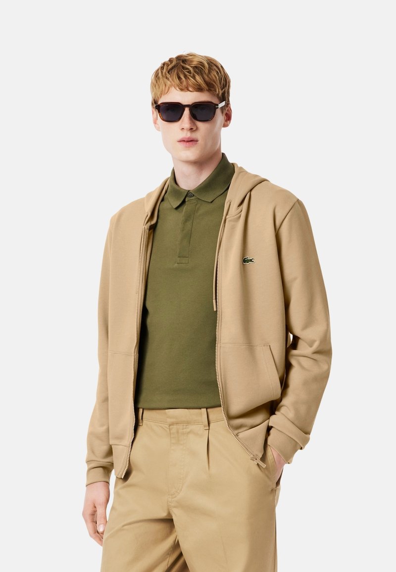 Sudadera con capucha de cremallera beige con bolsillos tipo canguro, usada sobre una camisa polo verde. El modelo también lleva gafas de sol oscuras. Los pantalones son de color beige claro.
