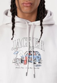 Valkoinen huppari, jossa on graafinen printti sinisestä autosta, sarjakuvamaisista rotiloista, vesihanan ja tekstin "JACKER" paksuilla kirjaimilla. Puuvillasekoitekangas.