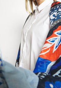 Sciarpa colorata a motivo appoggiata su una camicia bianca con bottoni. Il tessuto presenta disegni blu, arancio e nero con elementi floreali.