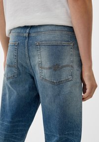 Vaqueros de denim azul desgastado con dos bolsillos traseros, acentos de costura visibles y detalles desgastados en los muslos. La cinturilla no es visible.
