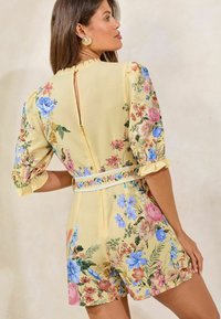 Floral romper in yellow fabric with puff sleeves, ruffled collar, and back zipper detail. Features colorful flower patterns throughout.