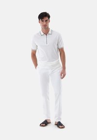 Weißes Polohemd mit schwarzem Besatz und Reißverschlusskragen. Kombiniert mit weißen Hosen und schwarzen Rutschsandalen. Glatte Textur, lässiges Design.