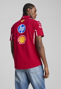 Polo roșu din bumbac, cu logo-uri ale sponsorilor (HP, Shell) pe spate, cu accente albe și un design clasic al gulerului.