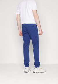 Blå stretchiga bomullbyxor med en avslappnad design, med bakfickor och slim fit, tillsammans med gråvita sportskor.