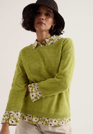 Žena oblečená v zelenom svetri cez kvetinovú blúzku, s béžovými nohavicami a tmavohnedým pleteným klobúkom, stojaca na neutrálnom pozadí.