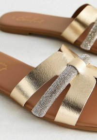 Sandali slides metallici dorati con una finitura texturizzata, caratterizzati da una cintura prominente decorata con strass. Sottopiede imbottito marrone.