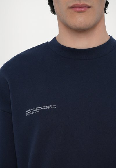 Mørkeblå sweatshirt lavet af økologisk bomuld, med en ribbet halsudskæring og en hvid trykt label med plejeinstruktioner på forsiden.