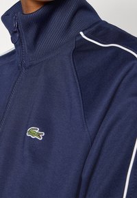 Chaqueta de Navy con cremallera y cuello alto, textura acanalada, detalles en blanco a lo largo de las mangas y un pequeño logo bordado de cocodrilo verde.