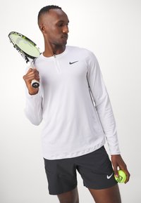 Camisa de rendimiento blanca de manga larga con cuello con cremallera y logo negro de Nike, combinada con pantalones cortos negros, sosteniendo una raqueta de tenis y una bola.