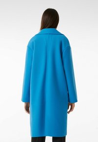 Lungo cappotto blu con un collo strutturato, maniche larghe e un taglio dritto. Il tessuto sembra liscio e spesso, adatto per il clima più fresco.