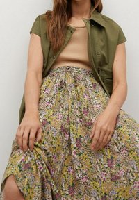 Lehká olivově zelená košile-jacket přes béžový top bez rukávů. Volná midi sukně v žluté barvě s květinovými vzory v růžové, fialové a zelené.