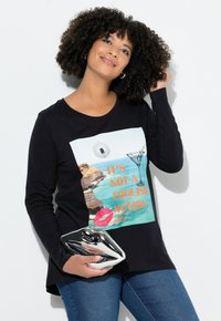 Langærmet sort t-shirt med et farverigt motiv, der inkluderer tekst, en cocktail og læber. Kombineret med blå jeans og en metallisk clutch.
