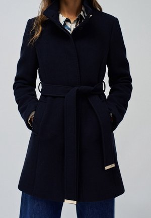 Femme portant un manteau en laine bleu marine foncé ceinturé par-dessus une chemise à motifs et un jean bleu, debout les mains dans les poches devant un fond uni.