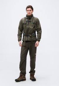 Άνδρας με εξωτερικό μπουφάν ελιάς, παντελόνι ταιριαστό, μποτάκια πεζοπορίας και γιλέκο με ζώνη ασφαλείας, στέκεται μπροστά σε απλό φόντο.