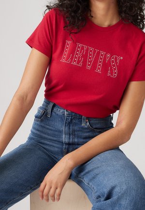 Czerwona bawełniana koszulka z logo "LEVI'S" z cyrkoniami, zestawiona z niebieskimi, wysokim stanem dżinsami, podkreślająca dopasowaną sylwetkę.