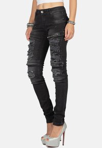 Jean skinny noir déchiré avec des détails usés, présentant une fermeture à bouton, cinq poches et un tissu texturé. Ornements en argent au niveau de la taille.