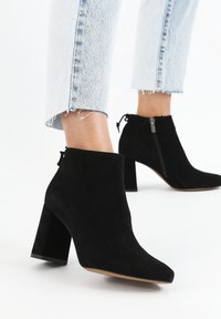 Stivaletti in suede nero alla caviglia con punta appuntita e tacco a blocco, dotati di una zip laterale e di una superficie liscia. Pantaloni in denim con risvolto visibili.