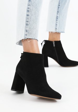 Stivaletti in suede nero alla caviglia con punta appuntita e tacco a blocco, dotati di una zip laterale e di una superficie liscia. Pantaloni in denim con risvolto visibili.
