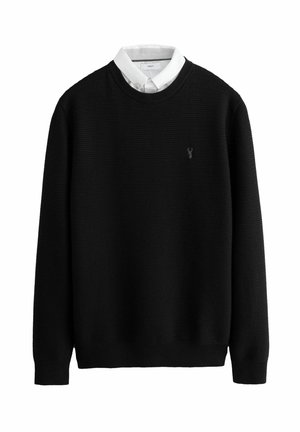 Crna pletiva pulover s rebrastom teksturom. Ima okrugli izrez i bijelu košulju s ovratnikom ispod. Mali logo izvezen na prsima.