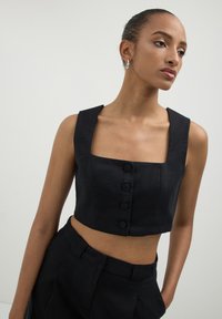 Donna che indossa un top a maniche corte nero con scollo quadrato e quattro bottoni, abbinato a pantaloni neri a vita alta, in piedi su uno sfondo neutro.