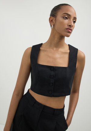 Donna che indossa un top a maniche corte nero con scollo quadrato e quattro bottoni, abbinato a pantaloni neri a vita alta, in piedi su uno sfondo neutro.