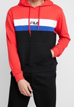 Man wearing red, white, blue, and black Fila hoodie with red drawstrings, right hand in pocket, left hand relaxed by side.