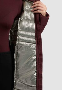 Giacca color marrone aperta che rivela una fodera interna lucida argento. Presenta dettagli trapuntati e una tasca nascosta all'interno. Texture liscia.