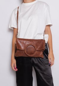Borsa a tracolla in pelle marrone con un accento frontale circolare, texture morbida e design pieghevole. Cinghia regolabile e dettagli in metallo argentato.