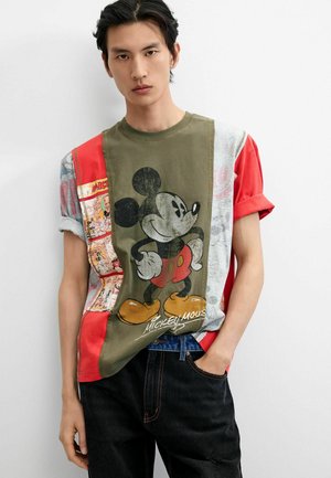 Młody mężczyzna o ciemnych włosach nosi wielokolorowy t-shirt z grafiką Myszki Miki i czarne jeansy, stoi na jednolitym białym tle.