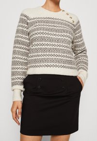 Maglione a maglia crema con motivi geometrici marroni e quattro bottoni dorati sulla spalla, abbinato a una mini gonna nera a vita alta.