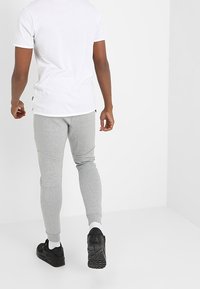 Person som går iväg iförd en vit t-shirt, ljusgrå joggingbyxor, vita strumpor och svarta sportskor mot en enkel vit bakgrund.