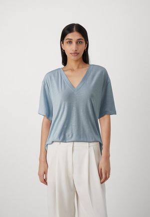 Maglietta azzurra chiara a maniche corte con scollatura a V in un tessuto morbido e liscio, caratterizzata da un fit rilassato e una texture sottile, abbinata a pantaloni bianchi larghi.