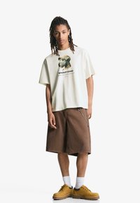 T-shirt crema a maniche corte con grafica di un cane e testo, abbinata a pantaloni corti marroni fino al ginocchio, scarpe gialle e calze bianche.