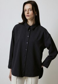 Black striped button-up shirt with a collar, loose fit, wide sleeves, and purple buttons. Smooth fabric with a subtle sheen.