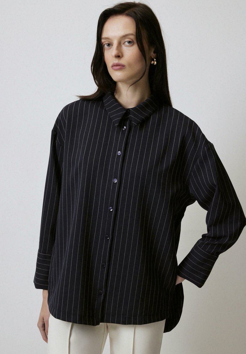 Black striped button-up shirt with a collar, loose fit, wide sleeves, and purple buttons. Smooth fabric with a subtle sheen.