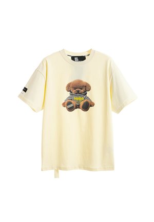 Joven vestido con camiseta crema con estampado de oso de peluche, pantalones cortos vaqueros azules con detalle de oso, calcetines blancos con estampado, y zapatos beige sin cordones.