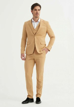 Costume beige en trois pièces avec un blazer ajusté, fermeture à un bouton unique et deux poches à l'avant. Le pantalon est slim-fit ; associé à des chaussures noires.