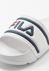 Ciabatta bianca con strisce blu navy e un prominente logo Fila nero e rosso sulla cinghia. Superficie texturizzata con design curvato alla base.
