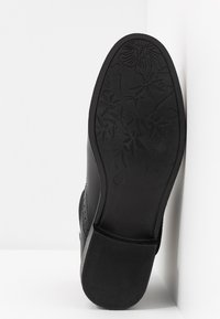 Scarpa nera con tomaia in pelle liscia, caratterizzata da dettagli traforati e suola in gomma. Motivo floreale intricato sulla superficie della suola.